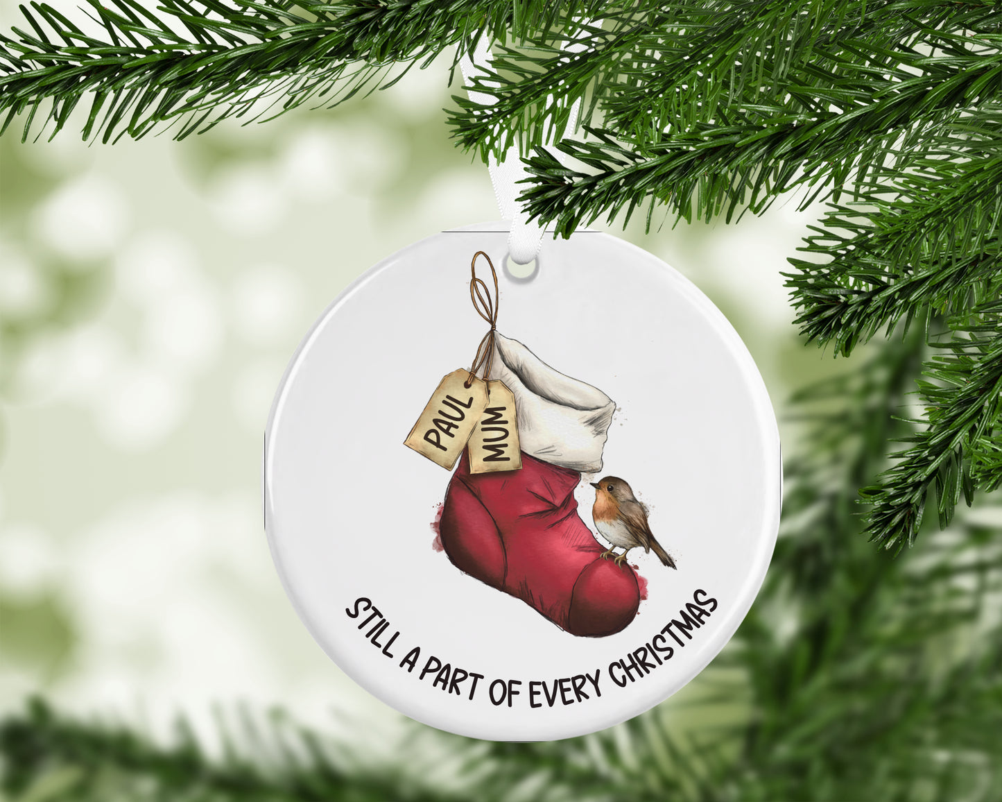 A round white Christmas ornament hanging from a green pine branch. The ornament features an illustration of a red stocking with a hanging tag that says 'PAUL MUM', a small robin bird, and the text 'STILL A PART OF EVERY CHRISTMAS'.